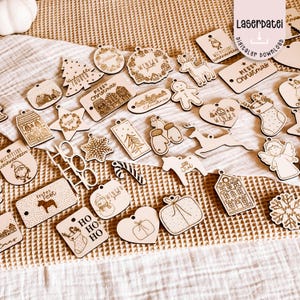 May include: A collection of laser-cut wooden Christmas ornaments in various shapes, including houses, stars, and reindeer. Some ornaments feature text like "Merry Christmas" and "Winter". The ornaments are light brown and have a rustic, handmade appearance.