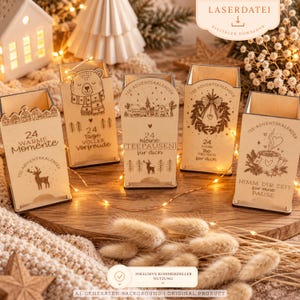 Könnte beinhalten: Fünf helle Holzboxen für Tee-Adventskalender mit lasergravierten Wintermotiven und deutschem Text. Jede Box zeigt einzigartige Designs wie einen Bären, ein Dorf, einen Kranz und eine Teetasse, wobei die Zahl 24 jeweils deutlich zu sehen ist.
