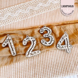 Può includere: Quattro ornamenti numerici in legno, da 1 a 4, con base bianca e disegni floreali neri. Ogni numero ha un piccolo foro nella parte superiore per essere appeso. I numeri sono esposti su un tessuto marrone.