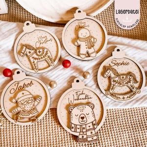 Puede incluir: Una colección de adornos navideños de madera con varios diseños. Los adornos incluyen un caballo, un hombre de jengibre, Papá Noel, un oso y un caballito balancín. Cada adorno tiene un lazo para colgar y está grabado con texto.