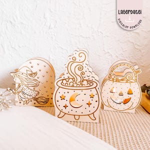Puede incluir: Decoraciones de Halloween de madera que representan una bruja en una escoba, un caldero con una luna creciente y una calabaza con cara. La bruja lleva un sombrero y un vestido. El caldero tiene estrellas y una luna creciente. La calabaza tiene una cara sonriente.