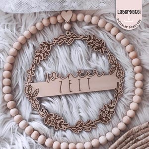 Op de afbeelding: Een houten krans met een bloemenontwerp en de woorden "Winter Zeit". De krans is omgeven door houten kralen en een klein houten hart. De tekst "Laserdatei digitaler download" staat in de rechterbovenhoek.