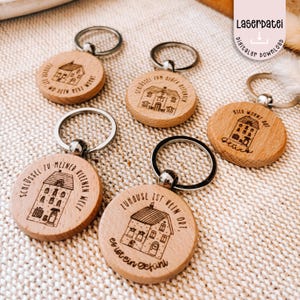 Puede incluir: Varios llaveros de madera con aros plateados. Cada llavero presenta un diseño de casa grabado con láser y texto en alemán. Los llaveros son redondos y están hechos de madera clara. El texto incluye frases como "Donde está tu corazón" y "El hogar no es un lugar."