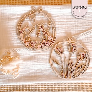 Pode incluir: Dois ornamentos de madeira com tema de primavera. Um apresenta a palavra "Spring" com desenhos florais, e o outro mostra flores de tulipa. Ambos são circulares com detalhes recortados intrincados e um laço no topo.