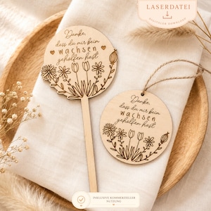 Può includere: Due segnaposto per piante in legno con disegni floreali e il testo tedesco "Danke, dass du mir beim wachsen geholfen hast." Un segnaposto è rotondo con uno spago, l'altro ha uno stelo. Lo sfondo include un piatto di legno e un panno bianco.