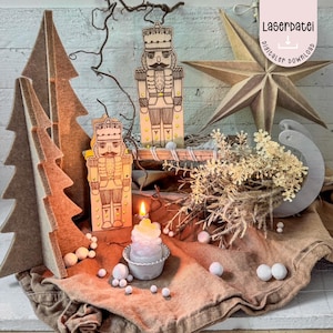May include: A festive holiday display featuring wooden nutcracker figures, a star, and felt Christmas trees. A lit candle sits in a small holder, surrounded by white decorative balls and dried flowers. The image includes the text "Laserdatei digitaler download".