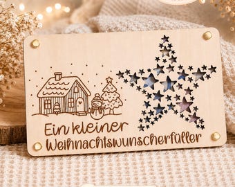 May include: A wooden Christmas decoration with a laser-cut design. It features a house, snowman, and Christmas tree on the left, and a star made of smaller stars on the right. The text "Ein kleiner Weihnachtswunscherfüller" is engraved below the house.