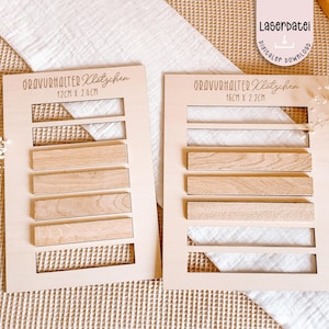 May include: Two wooden display boards with rectangular cutouts and wooden blocks. The boards have the text "GRAVURHALTER Klotzchen" and measurements of 12cm x 24cm and 16cm x 22cm. The boards are light-colored with a natural wood grain.