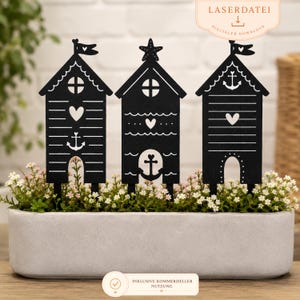 May include: Three black laser-cut plant stakes shaped like beach huts, each featuring nautical motifs like anchors, hearts, and waves. The stakes are inserted into a rectangular gray planter filled with small white flowers. Text reads LASERDATEI.
