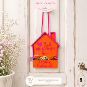 Könnte beinhalten: Ein hausförmiger Wandbehang mit leuchtend orangefarbener Basis und pinkfarbenem Rand, der den Schriftzug "-lich WILLKOMMEN bei familie Hedrich" in erhabenen pinken Buchstaben, ein pinkes Herz und ein kleines Blumenfach enthält.