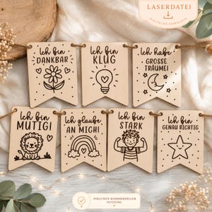 Könnte beinhalten: Ein Set aus sieben hellen Holzanhängern an einer Schnur, jeweils mit schwarzen Gravuren versehen, darunter eine Blume, eine Glühbirne, ein Mond, ein Löwe, ein Regenbogen, ein Kind beim Muskelzeigen und ein Stern mit positiven deutschen Sprüchen.