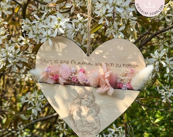May include: A wooden heart-shaped ornament with a cutout design of a mother and child. The ornament has a slot for dried flowers and the text "Glück ist eine Mama wie dich zu haben" is engraved on the wood.