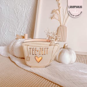Puede incluir: Un portavelas de madera con forma de taza de té con las palabras "TEE-ZEIT" y un recorte de corazón. El portavelas está rodeado de calabazas decorativas y un jarrón con flores secas. La imagen también incluye una etiqueta que dice "Laserdatei digitaler download".