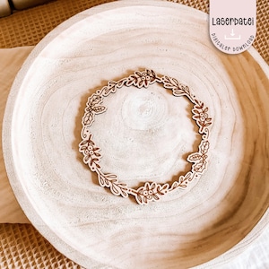 Puede incluir: Una corona de madera cortada con láser con un diseño botánico con hojas y bayas. La corona es de color marrón claro y se encuentra sobre un cuenco de madera de color claro. La imagen incluye el texto "Laserdatei digitaler download".