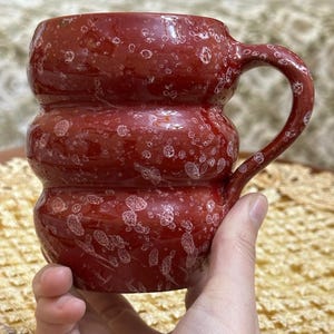 Puede incluir: Una taza de cerámica roja hecha a mano con un diseño texturizado único. La taza tiene una forma cilíndrica con tres secciones apiladas y un asa curva. Las motas blancas añaden interés visual al esmalte rojo.