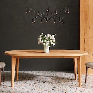 Könnte beinhalten: Ein ovaler Esstisch aus hellem Holz mit einer Vase mit weißen Blumen in der Mitte. Zwei Stühle mit hellgrauer Polsterung und dunklen Holzbeinen flankieren den Tisch. Über dem Tisch hängt ein schwarzer Kronleuchter.