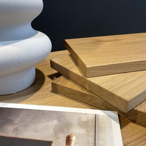 Op de afbeelding: Een stapel van drie lichte eikenhouten planken met een natuurlijk nerfpatroon. Een witte, gebogen decoratieve vaas staat links. Een afgedrukte afbeelding met een grijs en bruin ontwerp staat op de voorgrond.