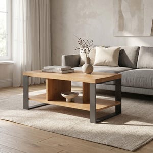 Könnte beinhalten: Ein rechteckiger Couchtisch aus hellem Holz mit einem unteren Regal und dunkelgrauen Metallbeinen. Der Tisch hat eine natürliche Holzoberfläche und enthält Bücher, eine Vase mit Zweigen und eine dekorative Schale. Der Tisch steht vor einem grauen Sofa.