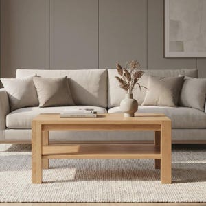 Könnte beinhalten: Ein rechteckiger Couchtisch aus hellem Eichenholz mit einem unteren Regal steht auf einem strukturierten Teppich. Hinter dem Tisch befindet sich ein beiges Sofa mit mehreren Kissen. Auf dem Tisch steht eine Vase mit getrockneten Blumen.