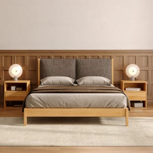 Op de afbeelding: Een houten bedframe met een donkergrijs gestoffeerd hoofdeinde, lichtgrijs beddengoed en een donkerbruine sprei. Twee nachtkastjes van licht hout flankeren het bed, elk voorzien van een bolvormige witte lamp met een centrale lichtbron.