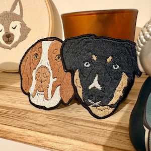 Peut inclure: Deux écussons brodés de chiens. L'un représente un chien marron et blanc avec un long museau. L'autre est un Rottweiler noir et fauve. Les deux écussons ont des bordures noires et sont sur une surface en bois.