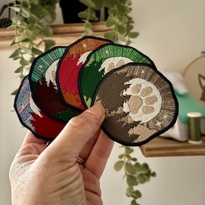 May include: A selection of embroidered patches with nature themes. Each circular patch has a black border and showcases a different landscape, such as trees, mountains, and a moon. The patches are in various colours, including red, green, and brown.