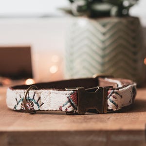 Peut inclure: Collier pour chien crème avec un motif tissé avec des accents rouges, bleus et verts. Le collier a un dos en cuir marron et une boucle et un anneau en métal de couleur bronze. Le collier repose sur une surface en bois.