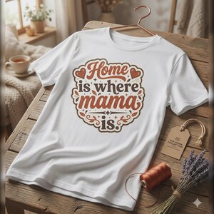 Peut inclure: T-shirt blanc avec l'inscription "Home is where mama is" dans un design marron et orange. Le t-shirt est présenté sur une surface en bois avec une étiquette indiquant "The Mama Collection". Une bobine de fil orange et un bouquet de lavande sont également présents.