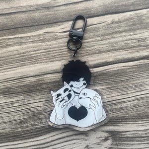 May include: A black and white acrylic keyring featuring a cartoon character holding two masks. The character has black hair and a heart on their shirt. The keyring has a black clasp.
