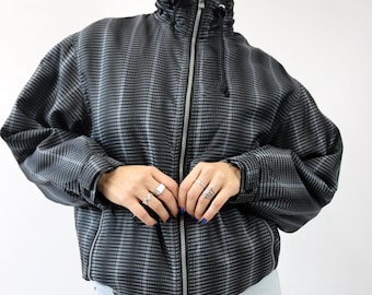 Könnte beinhalten: Eine schwarz-graue Jacke mit Karomuster, hohem Kragen und durchgehendem Reißverschluss. Die Jacke hat lange Ärmel und eine taillierte Passform. Die Person trägt eine hellblaue Jeans.