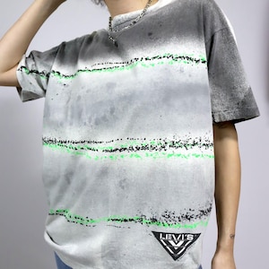 Può includere: Maglietta bianca con stampa astratta in nero e verde. La maglietta ha un logo Levi's nell'angolo in basso a destra.