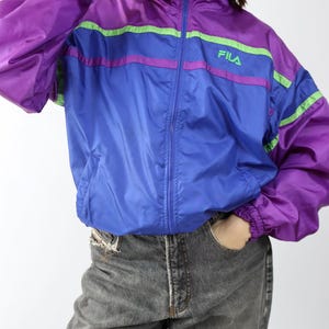 May include: A vintage Fila track jacket in purple and blue with green and pink stripes. The jacket has a high collar, a full zipper, and the Fila logo on the chest. The sleeves are long and the cuffs are elasticated.