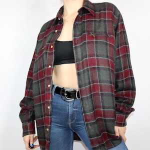 Puede incluir: Una camisa de franela a cuadros rojos y verdes con cierre de botones y mangas largas. La camisa está desabrochada y se lleva sobre un top negro sin mangas.