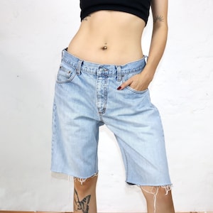 Könnte beinhalten: Hellblaue Jeansshorts mit rohem Saum. Die Shorts haben einen Knopfverschluss und Taschen. Der Denim ist verwaschen, was ihm einen Vintage-Look verleiht. Die Shorts sind knielang.