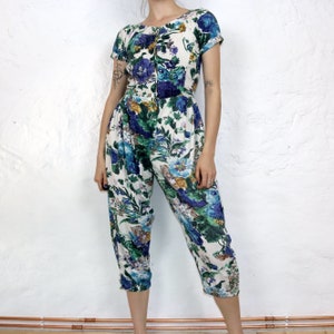 May include: A white jumpsuit with a floral print in shades of blue, green, and yellow. The jumpsuit has short sleeves and a scoop neck.