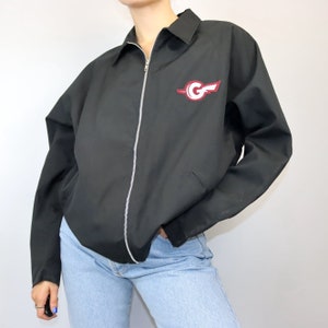 Könnte beinhalten: Schwarze Reißverschlussjacke mit einem roten und weißen Logo, auf dem "G" auf der linken Brust steht.