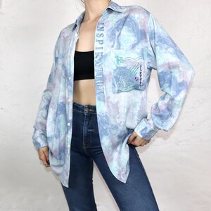 Op de afbeelding: Een lichtblauw overhemd met knopen en een tie-dye patroon. Het woord "Inspiration" staat verticaal gedrukt aan de linkerkant van het overhemd. Het overhemd heeft een grote zak aan de linkerkant met een grafisch ontwerp en het woord "Reizen" erop.