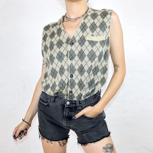 Könnte beinhalten: Eine ärmellose beige und schwarze Argyle-Weste mit Knopfleiste und einer kleinen Tasche auf der linken Seite.