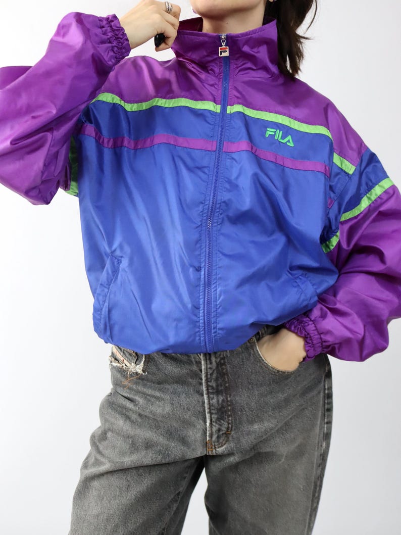 May include: A vintage Fila track jacket in purple and blue with green and pink stripes. The jacket has a high collar, a full zipper, and the Fila logo on the chest. The sleeves are long and the cuffs are elasticated.