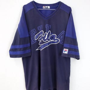 May include: Navy blue Fila V-neck t-shirt with blue sleeves and white Fila logo. The shirt has a classic athletic design with blue stripes on the sleeves and a small Fila logo patch.