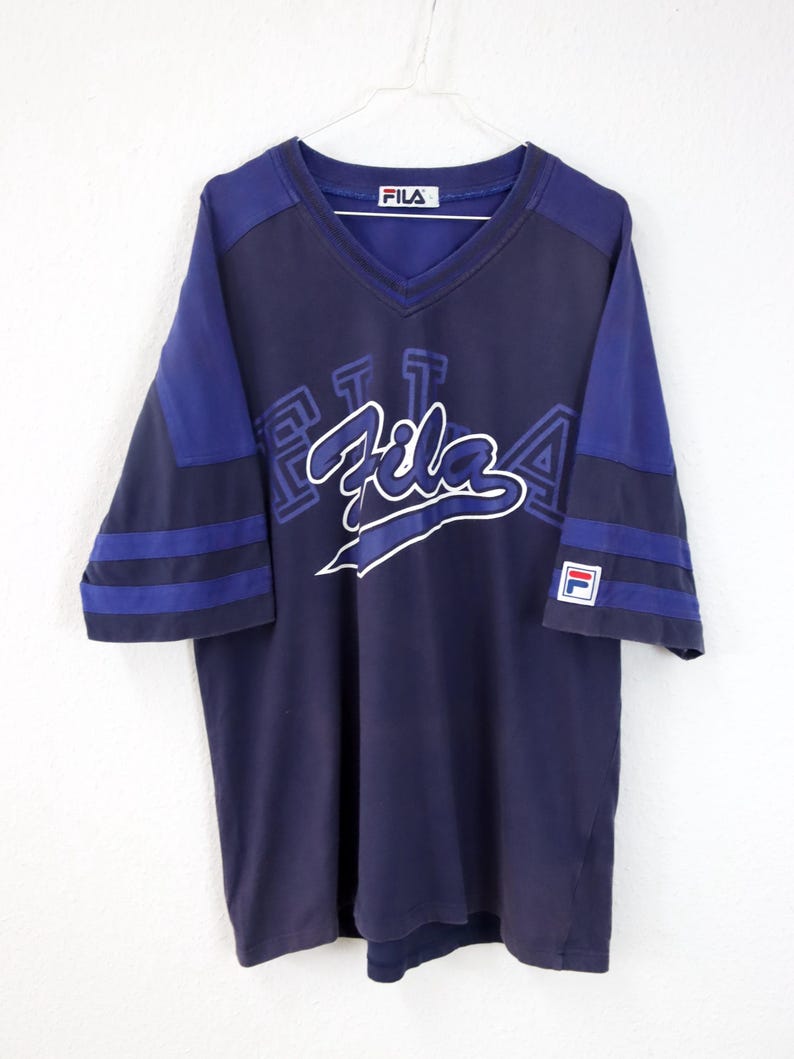 May include: Navy blue Fila V-neck t-shirt with blue sleeves and white Fila logo. The shirt has a classic athletic design with blue stripes on the sleeves and a small Fila logo patch.