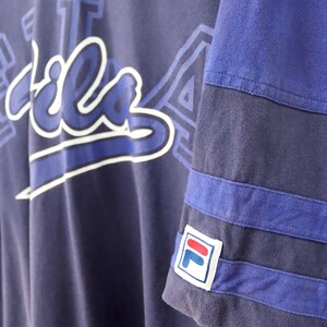 May include: Navy blue Fila t-shirt with white and blue lettering. The sleeve features blue stripes and the Fila logo. The shirt has a V-neck design and a relaxed fit.