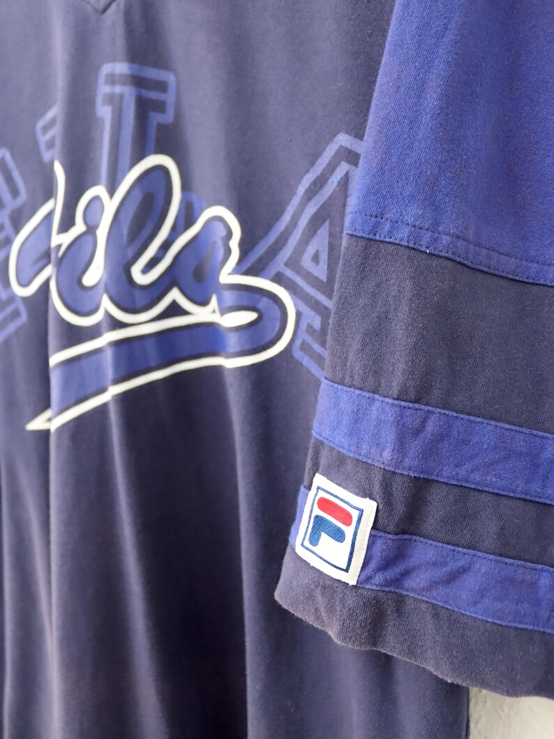 May include: Navy blue Fila t-shirt with white and blue lettering. The sleeve features blue stripes and the Fila logo. The shirt has a V-neck design and a relaxed fit.