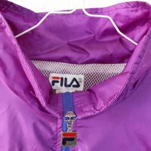 May include: A vintage purple Fila track jacket with a blue and green stripe across the chest. The jacket has a full zipper and the Fila logo is visible on the chest. The jacket is hanging on a white hanger.