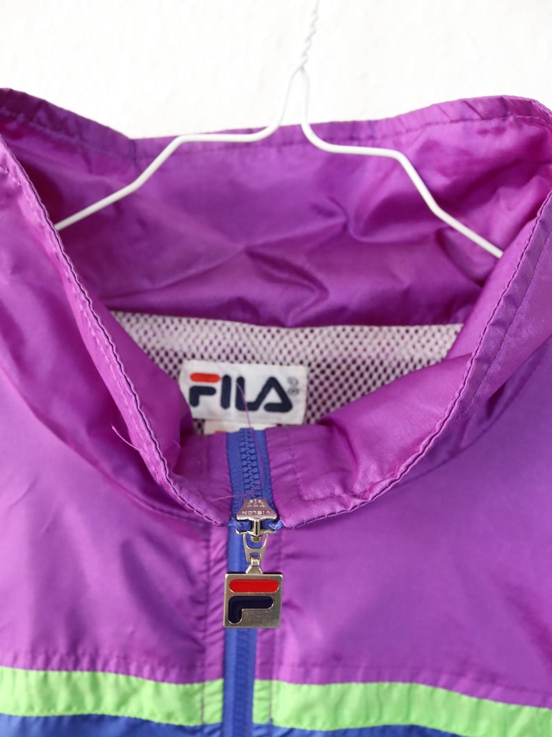 May include: A vintage purple Fila track jacket with a blue and green stripe across the chest. The jacket has a full zipper and the Fila logo is visible on the chest. The jacket is hanging on a white hanger.