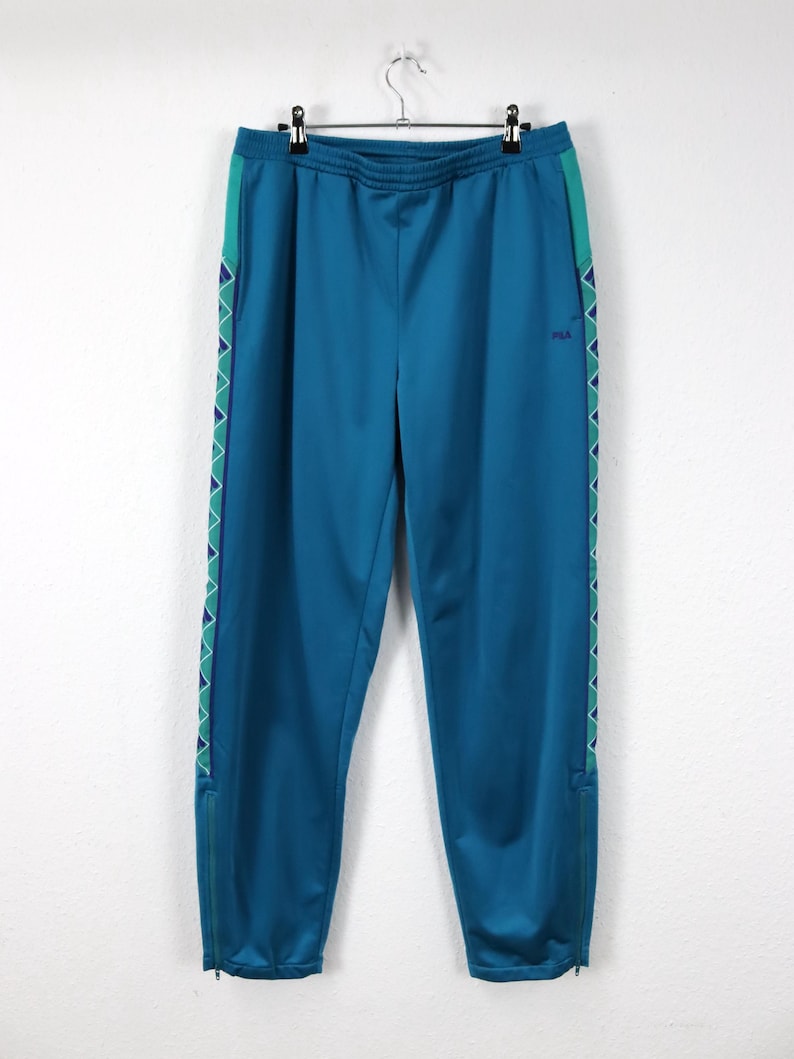 Puede incluir: Pantalones deportivos de color turquesa con cintura con cord&oacute;n y rayas laterales. Los pantalones tienen un patr&oacute;n geom&eacute;trico turquesa y morado en los laterales y un peque&ntilde;o logotipo de Fila. Los pantalones tienen cremalleras en los tobillos.