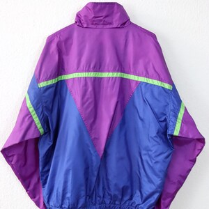 May include: A vintage windbreaker jacket in purple, blue, and green. The jacket has a high collar and a V-shaped design on the front. The sleeves and lower hem are elasticized. The jacket is hanging on a white background.