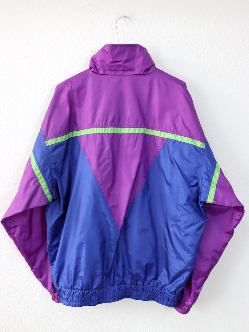 May include: A vintage windbreaker jacket in purple, blue, and green. The jacket has a high collar and a V-shaped design on the front. The sleeves and lower hem are elasticized. The jacket is hanging on a white background.