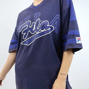 May include: Navy blue Fila t-shirt with a V-neck and short sleeves. The shirt features the Fila logo in white and blue across the chest, with blue stripes on the sleeves. A small Fila logo patch is on the sleeve.