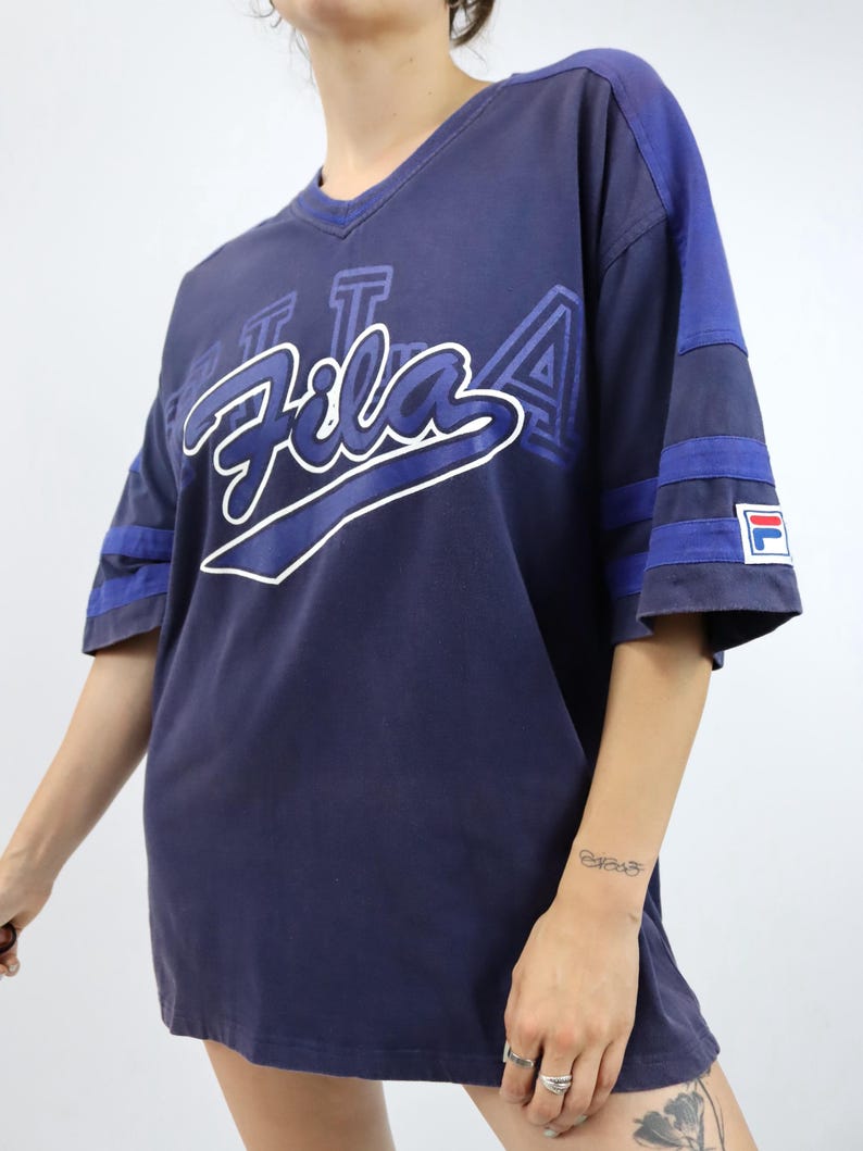 May include: Navy blue Fila t-shirt with a V-neck and short sleeves. The shirt features the Fila logo in white and blue across the chest, with blue stripes on the sleeves. A small Fila logo patch is on the sleeve.